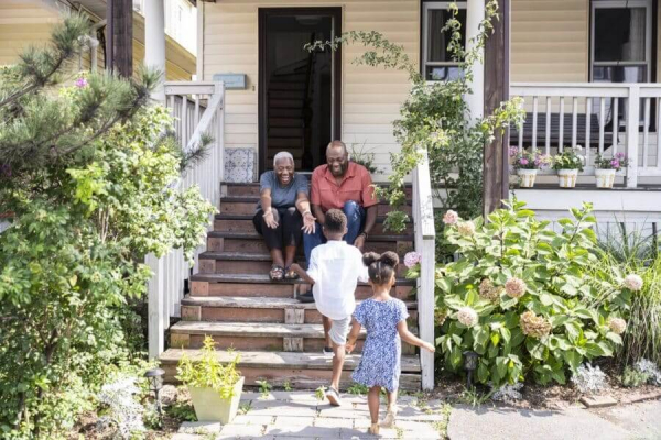 Full length view from behind 4 and 7 year old siblings as they run toward grandparents sitting on front porch steps, arms outstretched in greeting.